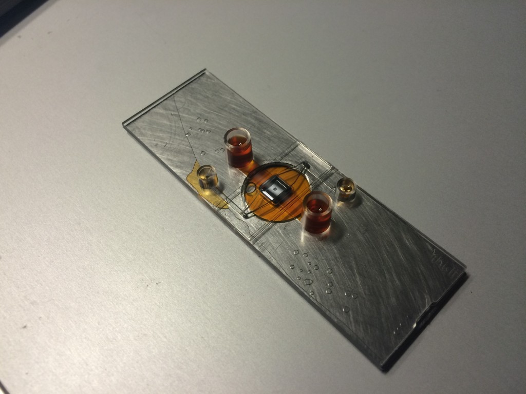 iBidi to MgF2 Floor bonding. The PDMS was spin coated at 1000 RPM for 100 s, cured for 1 hr at 50C on a hotplate. This thickness was enough to close off the top channel. The PDMS bonding between the brushed metal and the plastic appears good. The liquid pools underneath instead of staying in the bottom channel due to the recession of the coverslip. It remains unknown how the MgF2 will bond at this point, but it should have sufficient contact area to prevent this flooding.